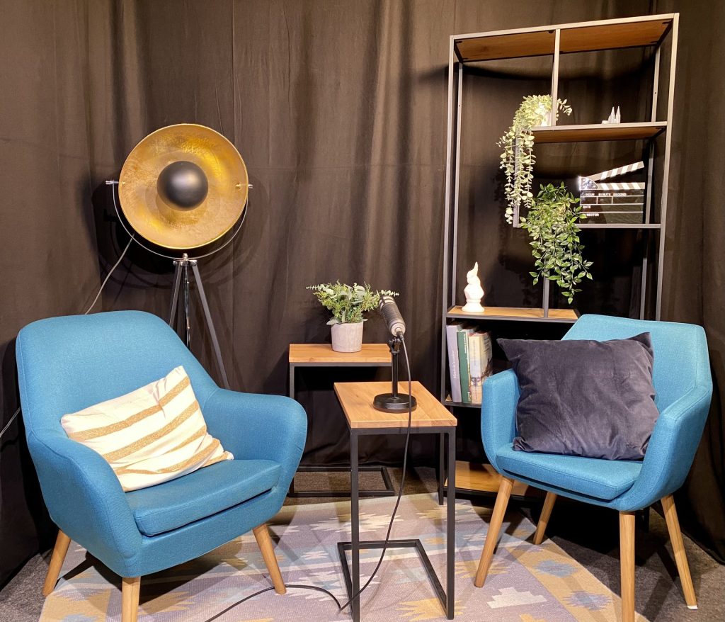 Das Social-Media-Studio der Stadtbibliothek Köln. Im Vordergrund zwei hellblaue Sessel und ein Tischchen mit einem Mikrofon. Dahinter ein Regal mit Deko, ein weiteres Tischchen und eine goldene Stehlampe aus Metall. Die Raumwand wird von einem schwarzen Vorhang komplett verdeckt.