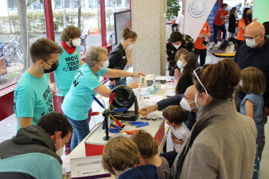 Ein dutzend Besucher*innen und vier Mitarbeitende stehen am Aktionsstand der Stadtbibliothek. Auf dem Tisch steht ein Experimentierkasten zum Thema Windenergie, der gerade von einem Jungen bedient wird. Andere Kinder probieren Ozobots und littleBits aus.