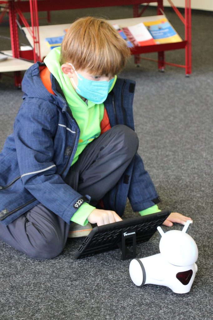 Ein Junge kniet vor einem Tablet-Computer und blickt auf einen kleinen, weißen, fahrbahren Roboter. Er bedient das Tablet, um den Roboter zu programmieren.